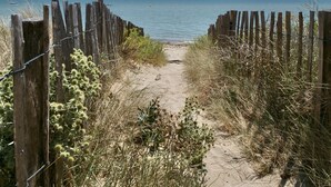 Ein sandiger Küstenpfad führt zum Meer, gesäumt von Holzzäunen und Vegetation.