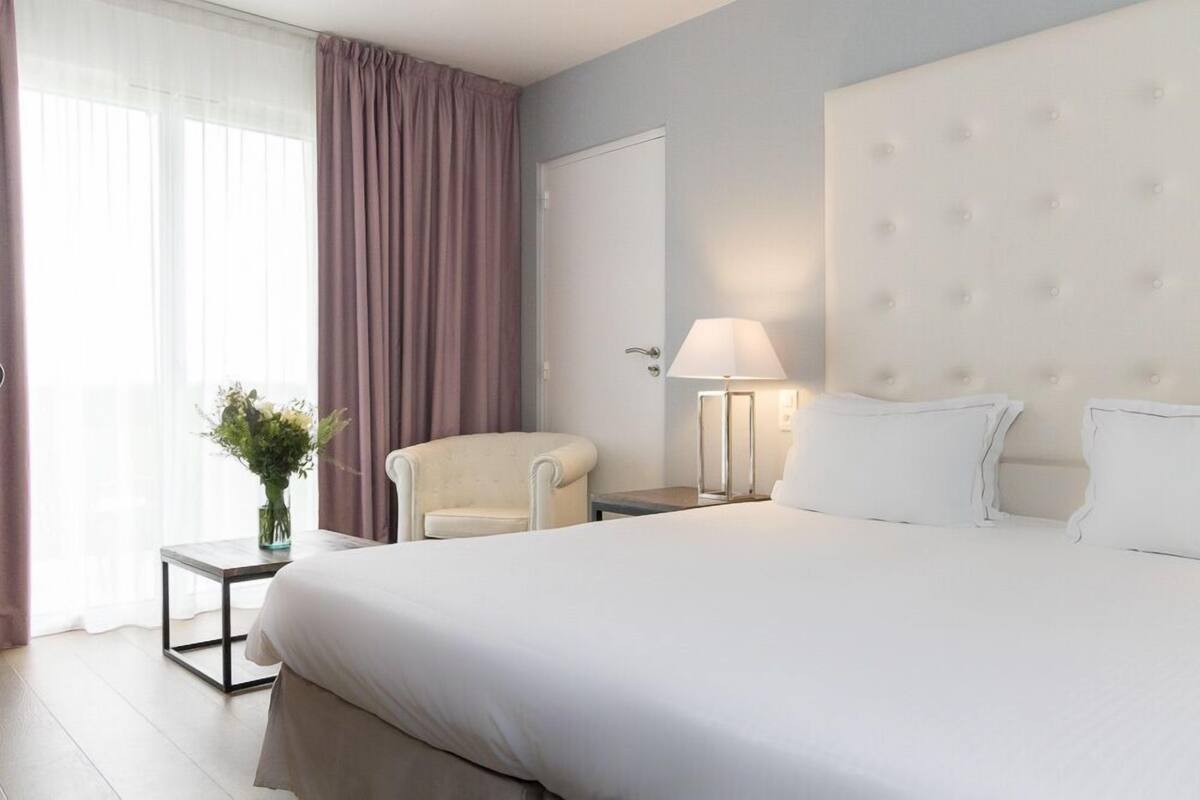 Une chambre d’hôtel avec un grand lit, une chaise, une table de chevet avec une lampe, un vase de fleurs et une fenêtre avec des rideaux.