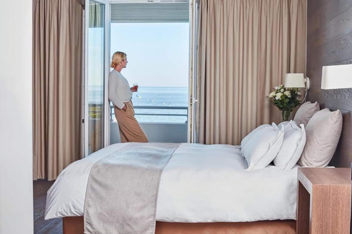 Une chambre à coucher avec un lit, des rideaux, une table de chevet, une lampe et une vue sur la mer par une porte ouverte.