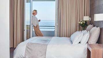 Une chambre à coucher avec un lit, des rideaux, une table de chevet, une lampe et une vue sur la mer par une porte ouverte.
