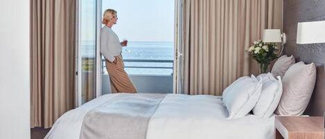 A bedroom with a bed, curtains, a nightstand, a lamp, and a view of the sea through an open door.