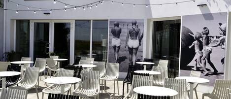 Un espacio al aire libre con mesas y sillas, un cartel grande con gente en la playa, y un letrero que dice "LA GUINGUETTE".