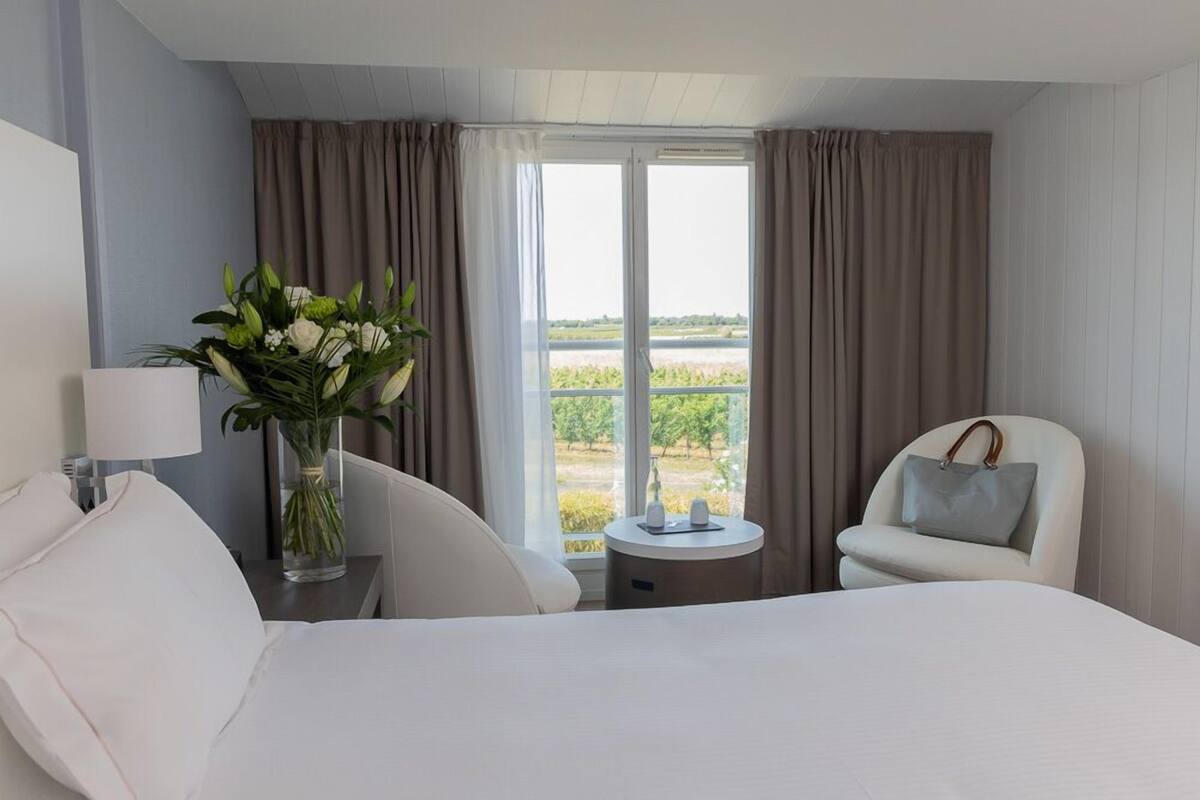 Une chambre d’hôtel avec un lit, une chaise, une petite table, un vase de fleurs et une vue sur un paysage verdoyant par la fenêtre.