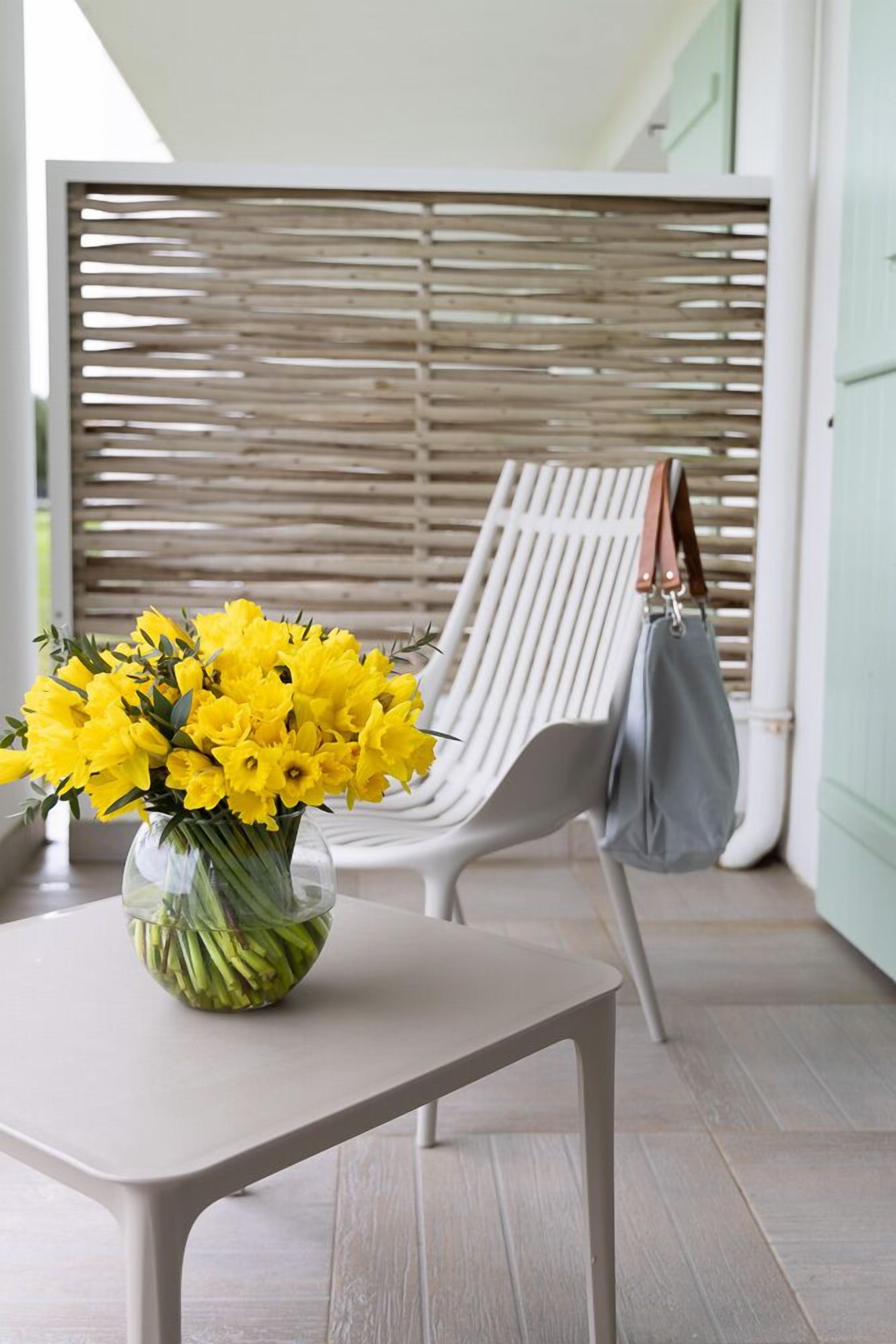 Una mesa blanca con un jarrón de flores amarillas, una silla blanca y un bolso gris, y una pared de listones de madera.