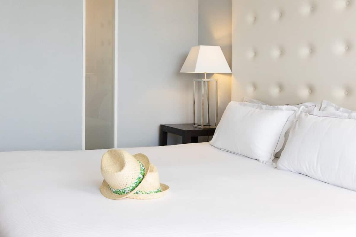 Un lit bien fait, avec du linge de lit blanc et des oreillers, une lampe de chevet et un chapeau de paille posé sur le lit.