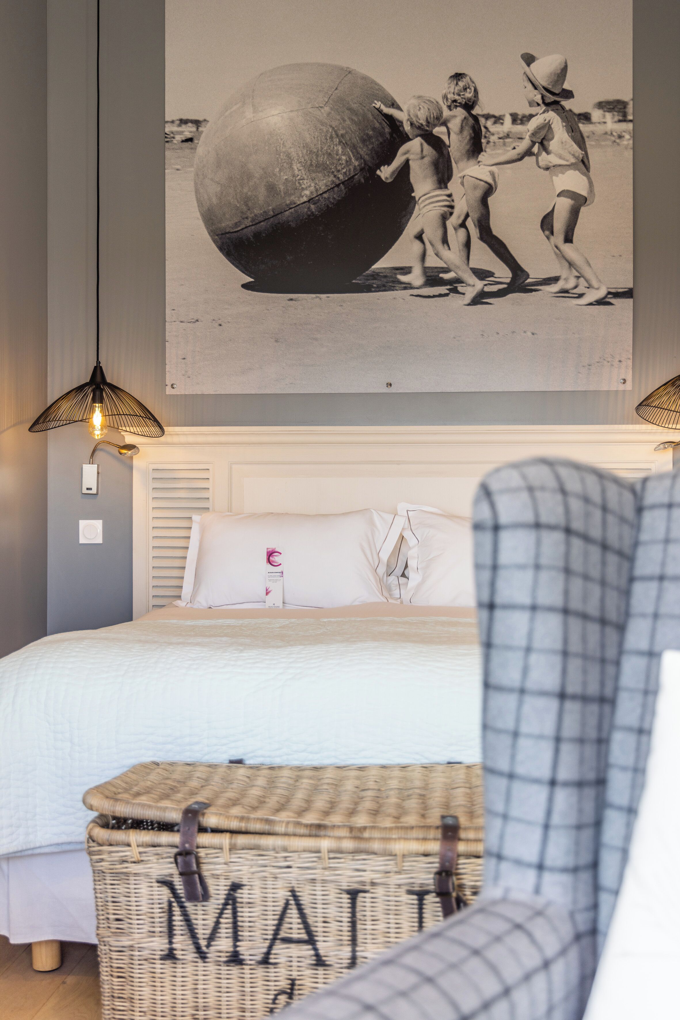 Un dormitorio con una cama, una cesta de mimbre que dice 'MAILING', una lámpara de pared y un cuadro enmarcado sobre la cama.