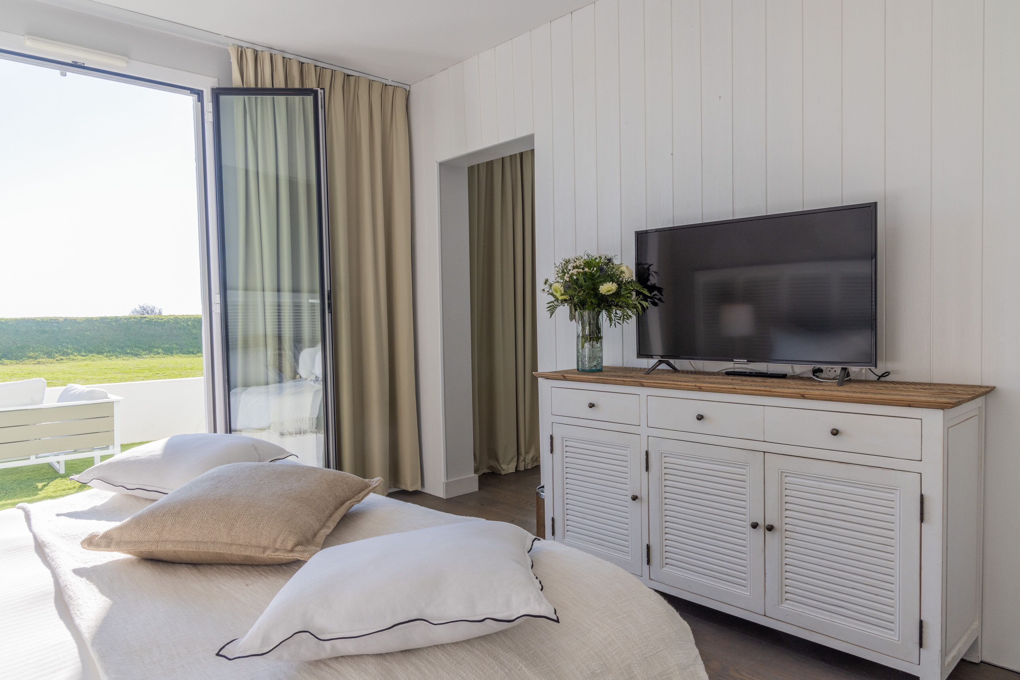 A bedroom with a large bed, a television on a wooden cabinet, and a view of the outdoors through a sliding door.