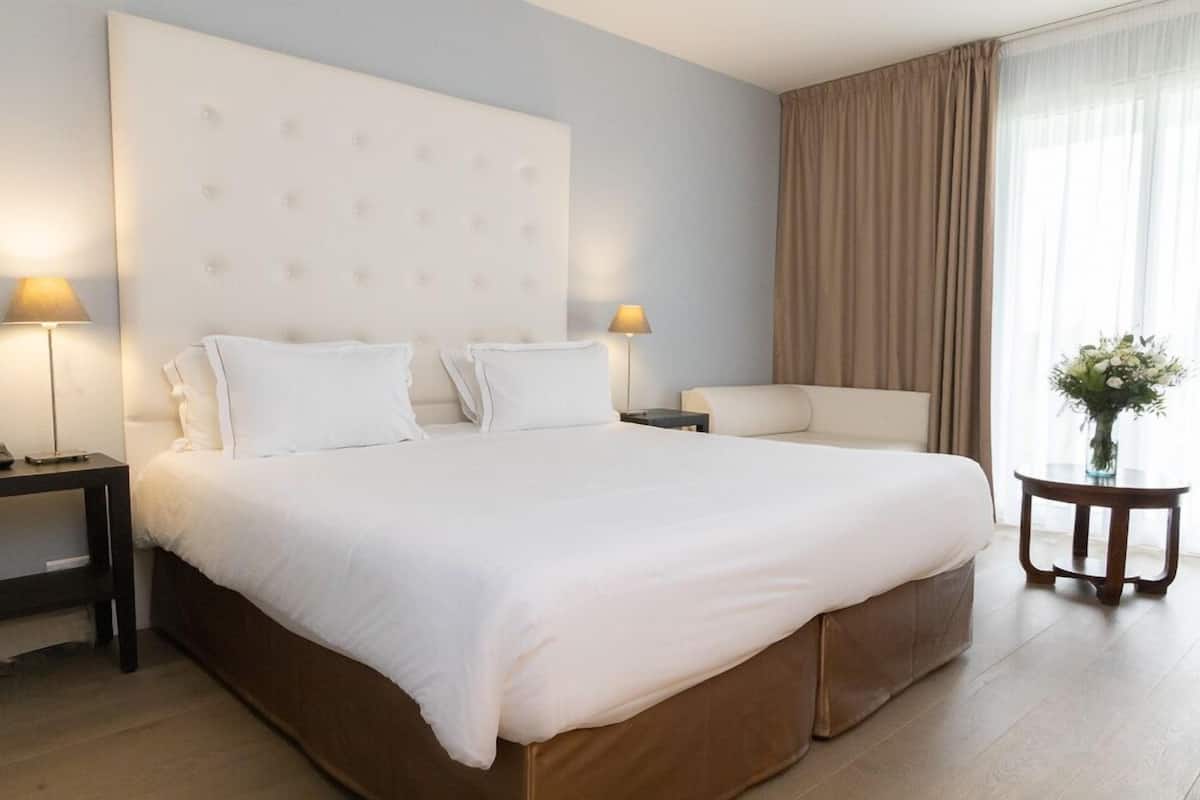 Une chambre d’hôtel avec un grand lit, deux tables de chevet, une petite table d’appoint, une chaise et une petite table ornée d’un vase de fleurs.