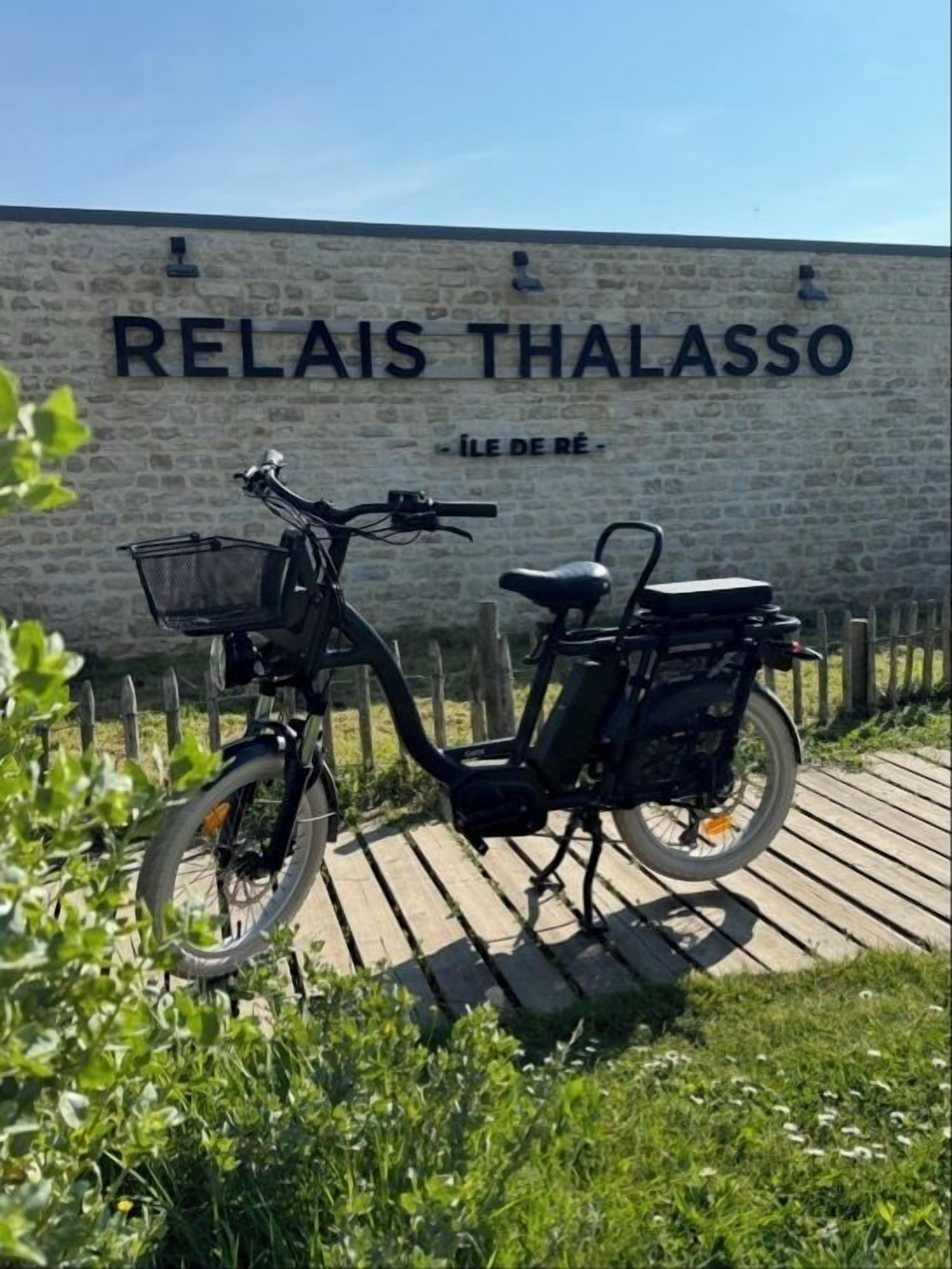 Uma bicicleta elétrica estacionada em frente a um muro com a sinalização 'RELAIS THALASSO' e 'ÎLE DE RÉ'.