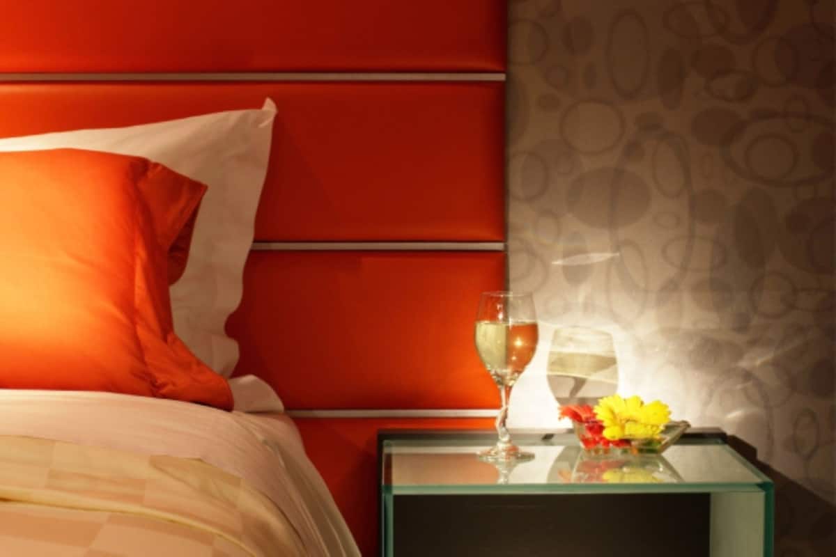 A bed with orange bedding, a glass of wine, and a vase with flowers on a glass table beside it.
