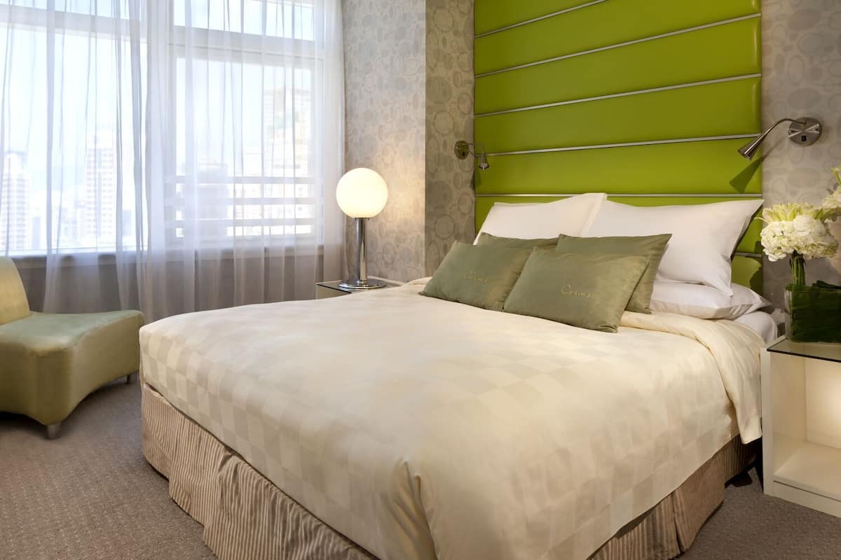 A hotel room with a large bed, a bedside table, a lamp, a chair, and a view of the city through the window.