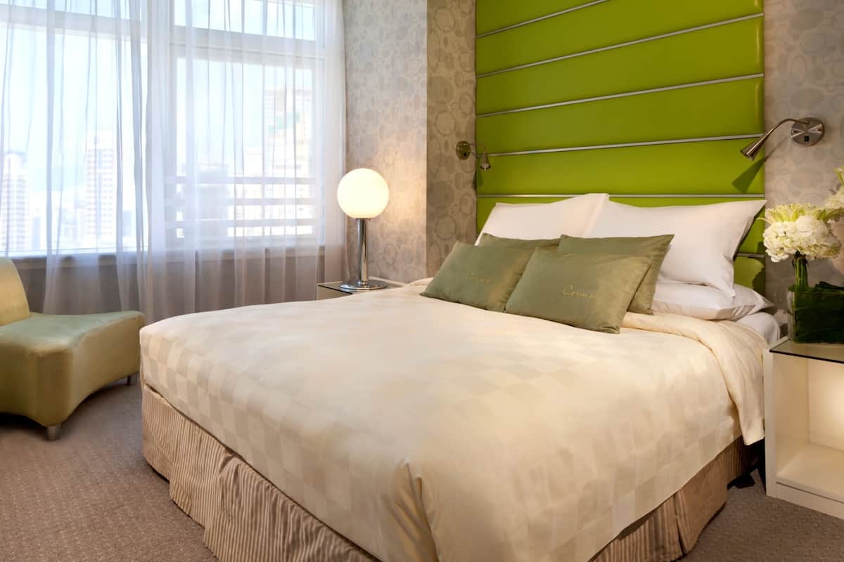 A hotel room with a large bed, a green headboard, a bedside table with a lamp, and a view of the city through sheer curtains.