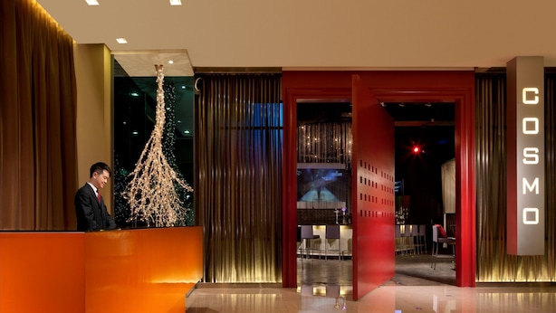 Zona de recepción de un hotel con diseño moderno, que incluye un mostrador de recepción, una lámpara de araña grande y una pantalla de televisión visible.