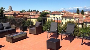 Varanda com móveis de vime, mesa de vidro e vista para uma torre histórica e edifícios.