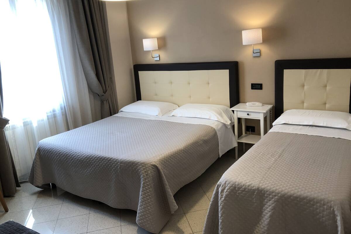 Habitación de hotel con dos camas, una mesita de noche con lámpara y una ventana con cortinas.