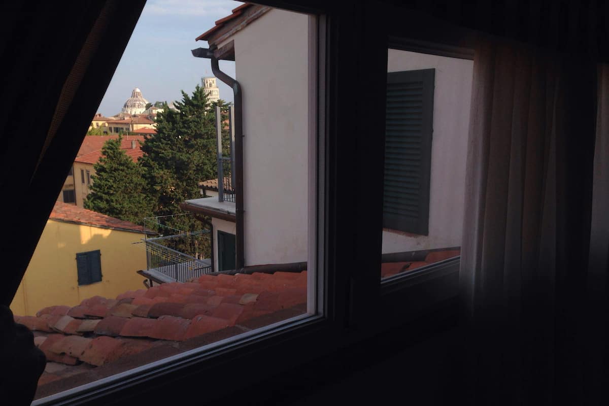 Una ventana con vista a un edificio con una cúpula y una torre, un árbol y un tejado de tejas rojas.