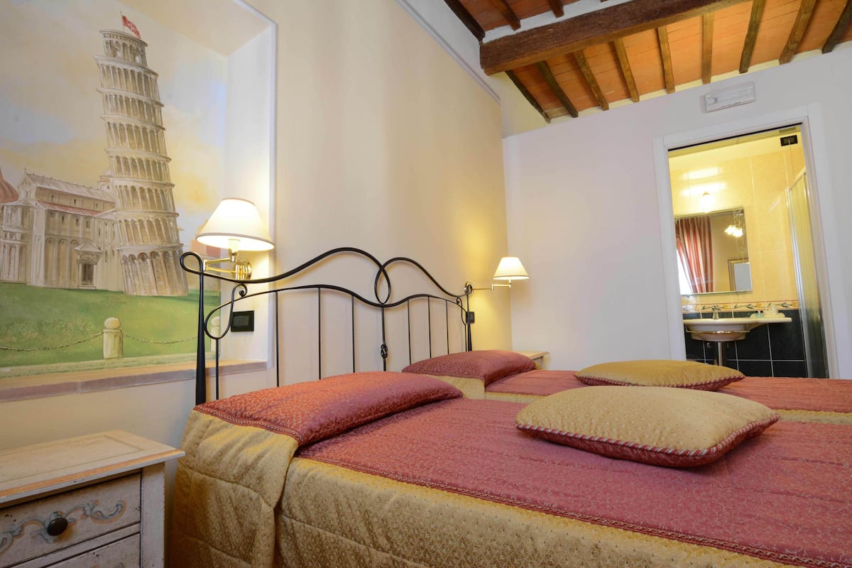 Una habitación de hotel con una cama, dos lámparas de noche, una mesita de noche, un aparador y un mural en la pared de la Torre de Pisa.