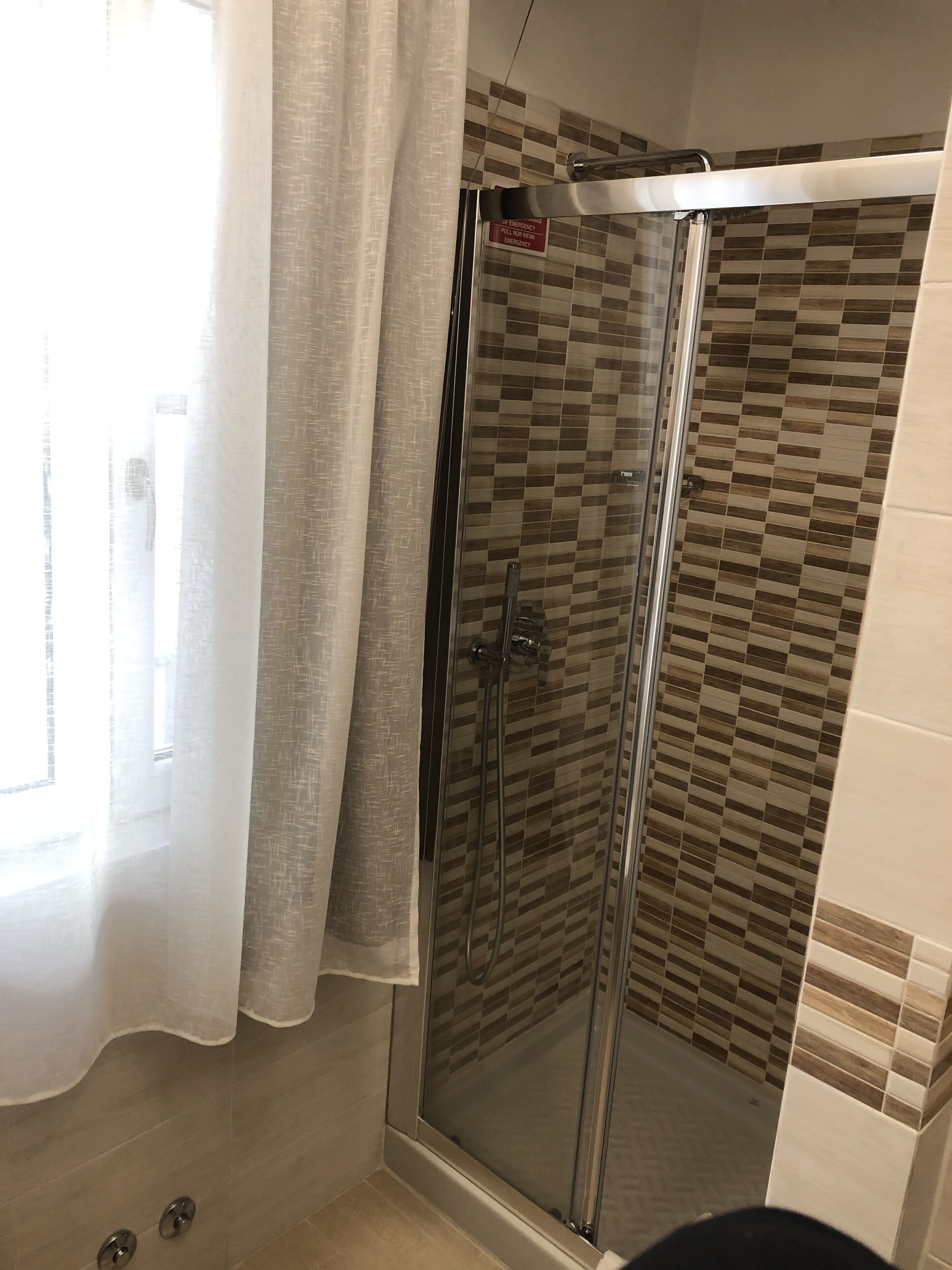 Une douche entièrementClose avec des murs recouverts de carreaux à motifs, une poignée de détresse bien visible et une fenêtre ornée d’un rideau transparent.