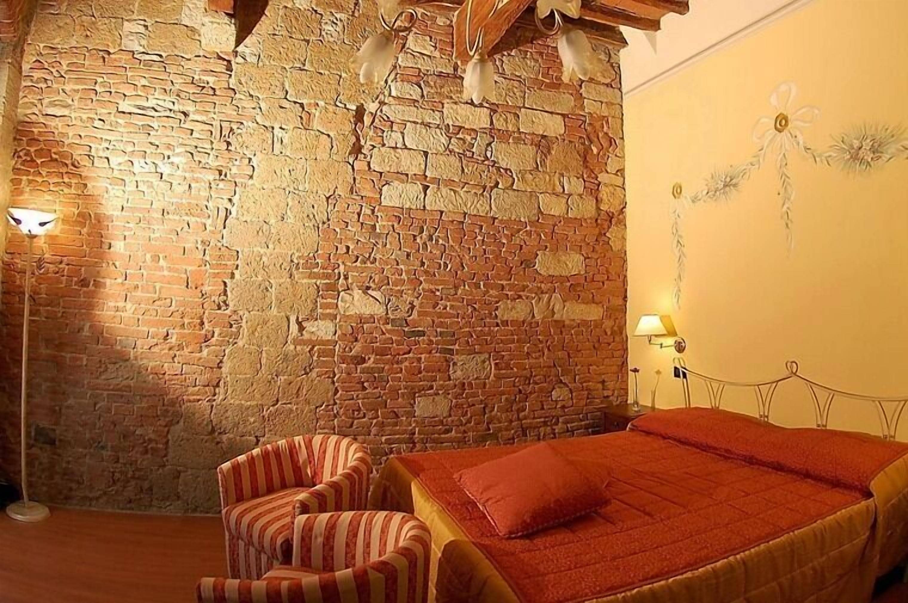 Une pièce avec un mur en briques, un lit recouvert d’une couverture rouge, des chaises à rayures et des lampes fixées au mur.