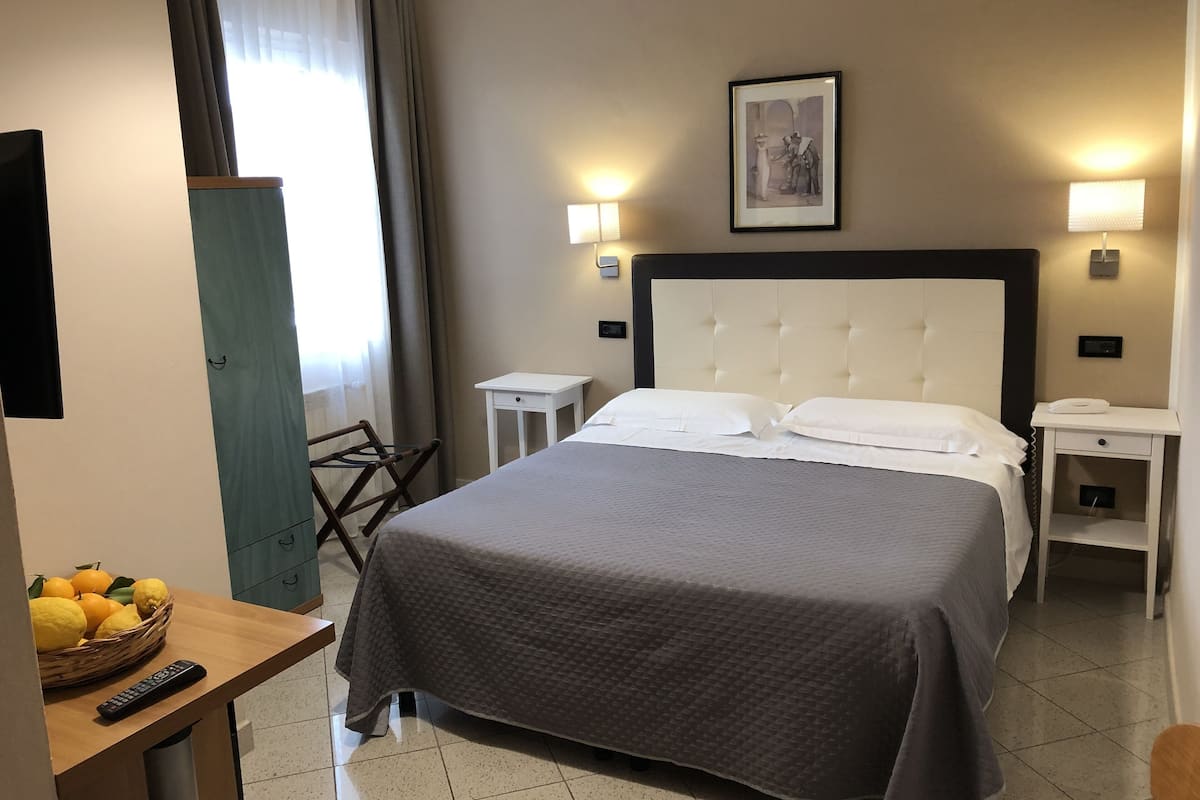 Habitación de hotel con una cama grande, mesitas de noche, una lámpara, un cuadro en la pared y una cesta de fruta sobre una mesita auxiliar.