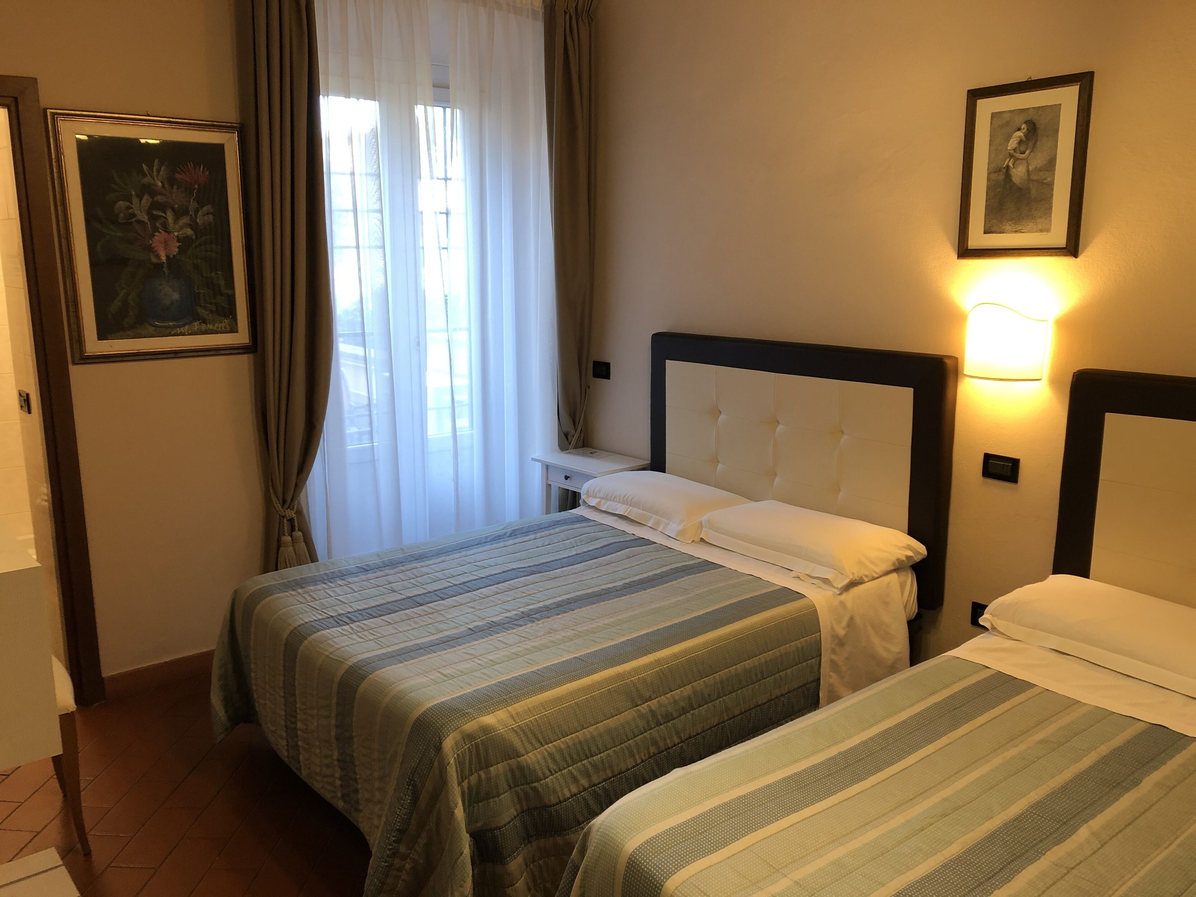 Een hotelkamer met twee bedden, een nachtkastje, een ingelijste foto en een raam met gordijnen.