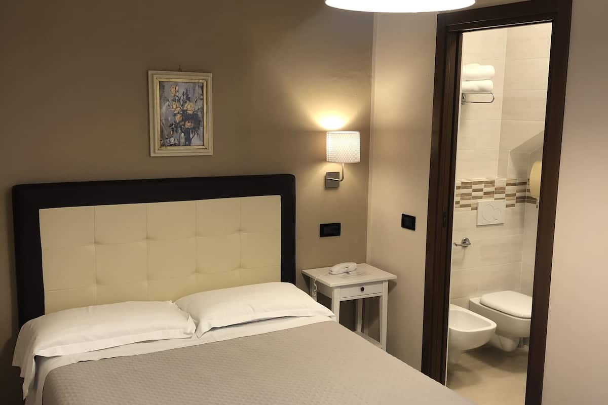 Una habitación de hotel con una cama grande, una mesita de noche con teléfono, una lámpara de pared y un baño visible a través de una puerta abierta.