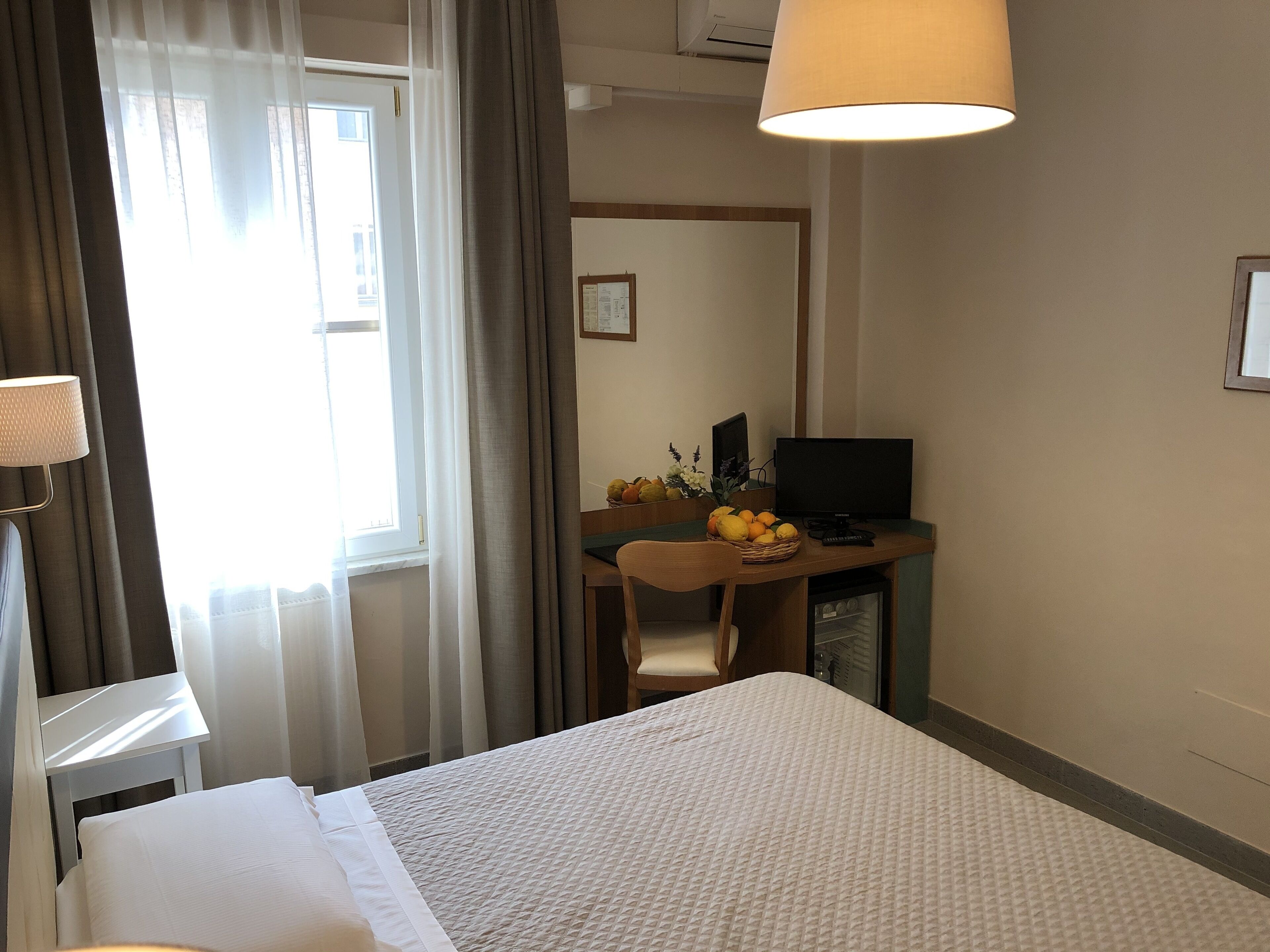 Habitación de hotel con cama, escritorio con computadora, silla, lámpara y ventana con cortinas.