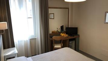 Una camera d'albergo con un letto, una scrivania con un computer, una sedia, una lampada e una finestra con tende.