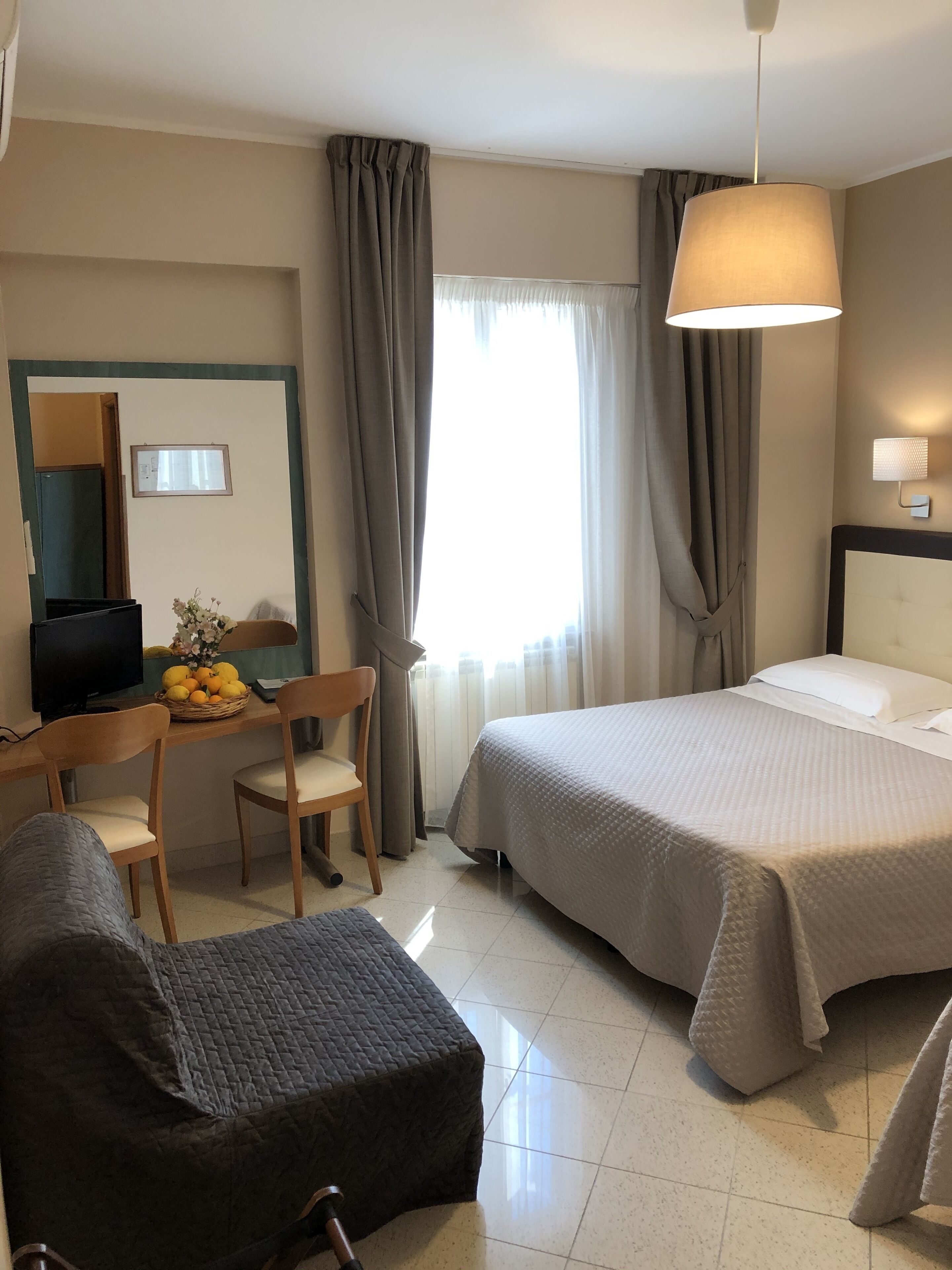 A hotel room with a bed, a desk with a computer, a chair, a television, and a window with curtains.