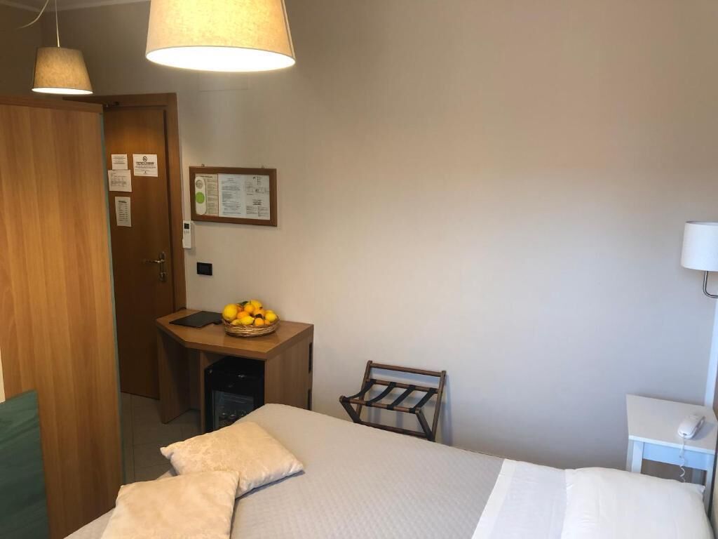 Una camera d'albergo con un letto, una scrivania con un cesto di frutta, una sedia, una lampada a parete e una porta.