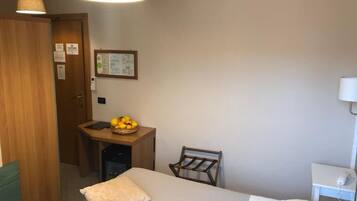 Una camera d'albergo con un letto, una scrivania con un cesto di frutta, una sedia, una lampada a parete e una porta.