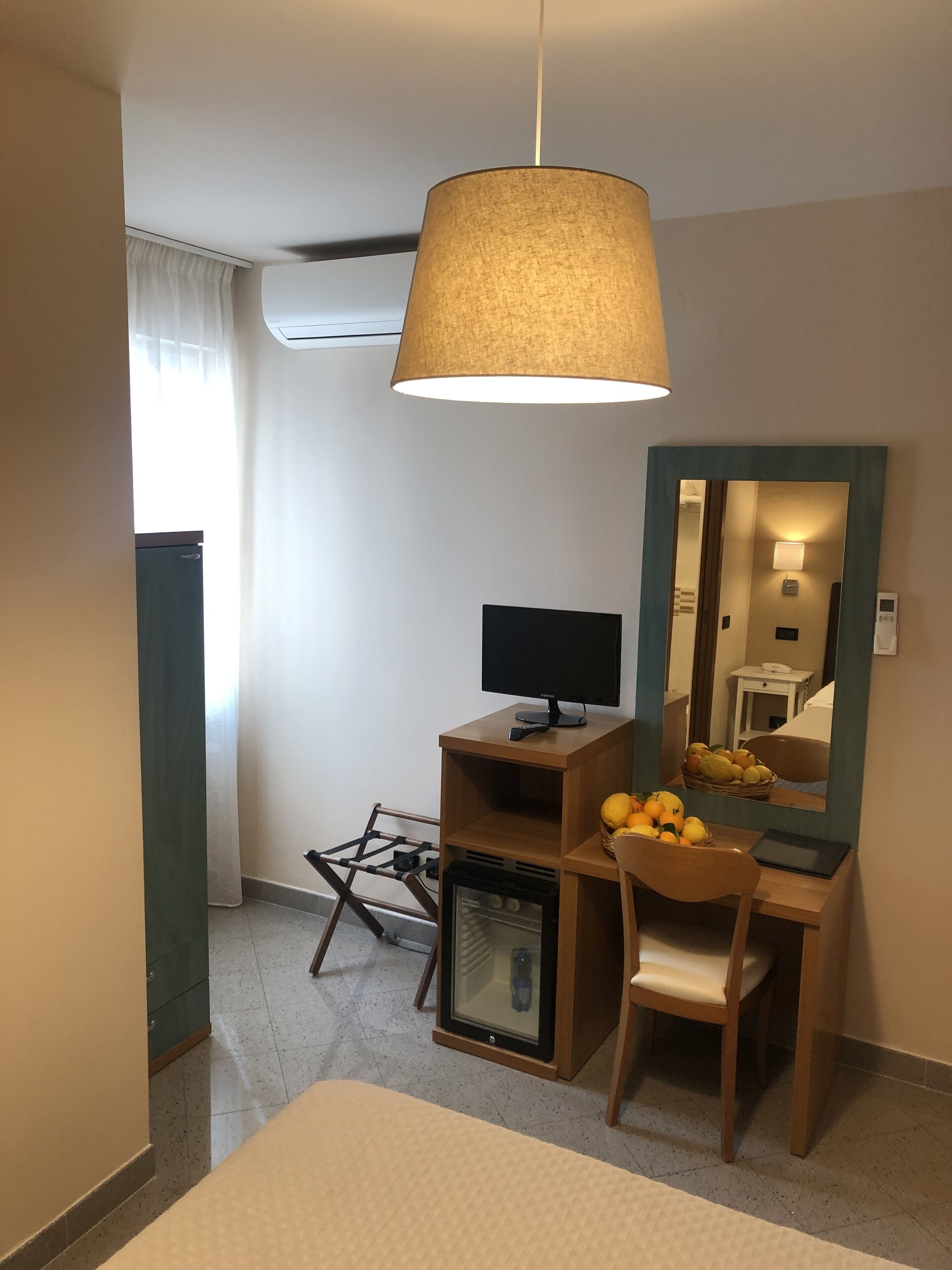 A hotel room with a bed, a desk with a computer, a mirror, a small refrigerator, and a hanging lamp.