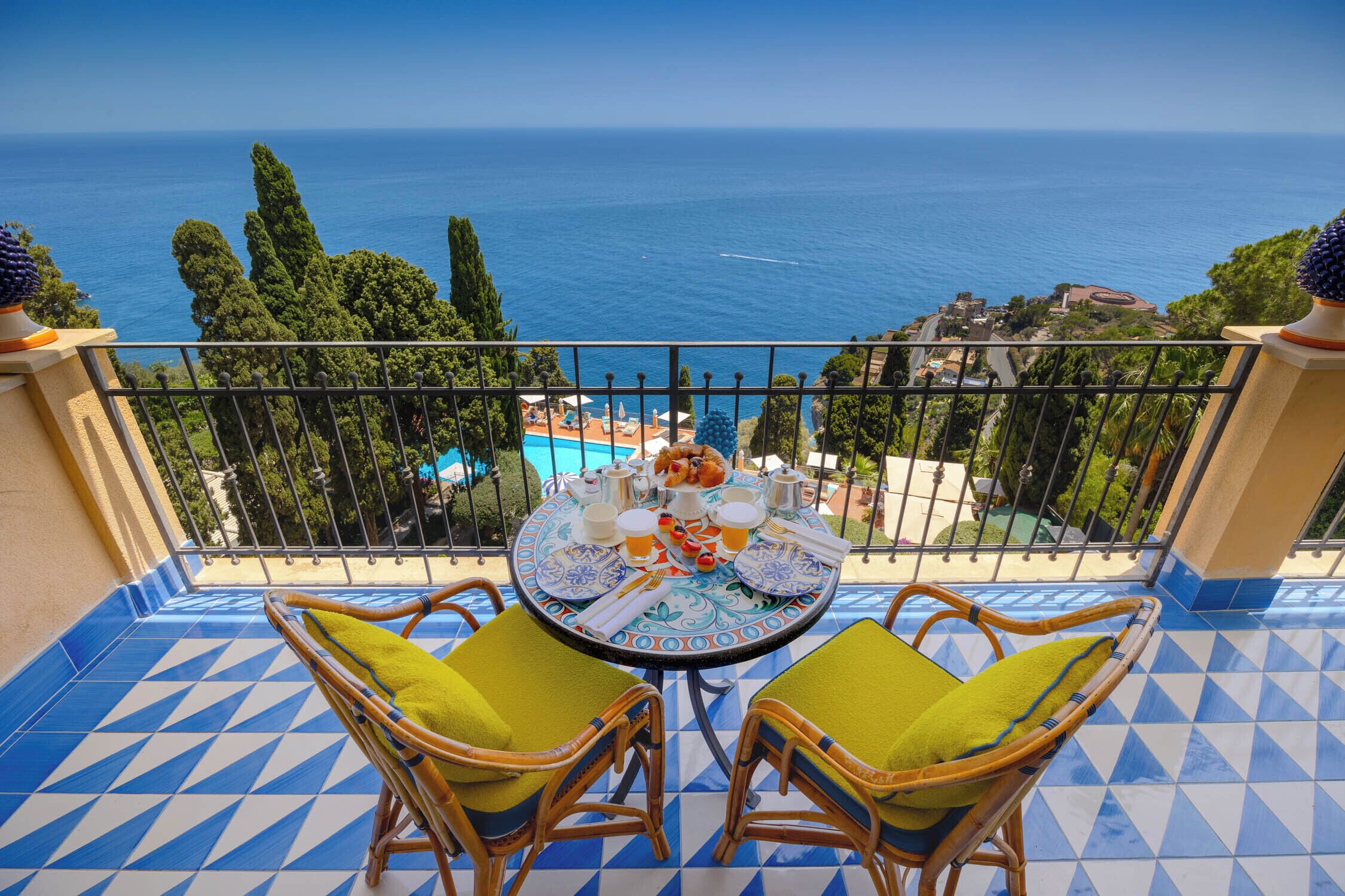 A balcony with a view of the sea, a round table with chairs, and a pool in the background.