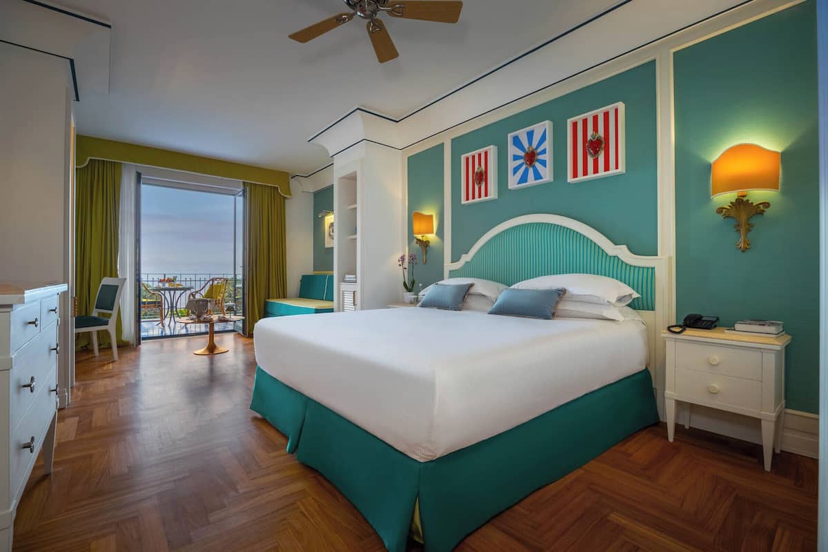 Habitació d'hotel amb un llit gran, un balcó amb taula i cadires, i vista al cel.