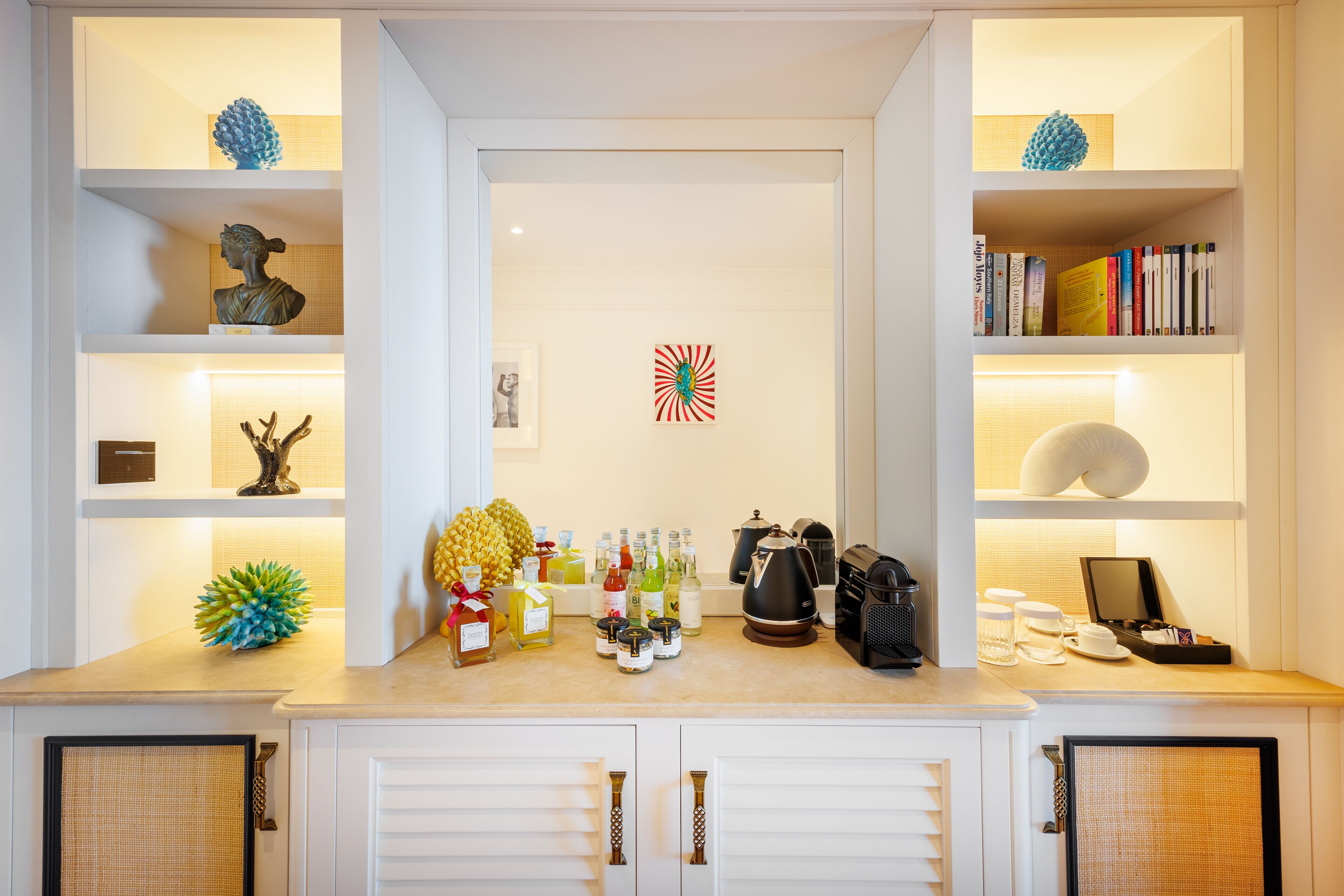 Una cocina bien organizada con una encimera que muestra varios objetos, entre ellos botellas, una cafetera y un frutero. Estantes a ambos lados exhiben objetos decorativos y libros.