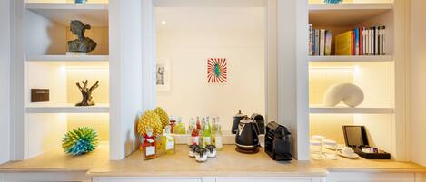 Una cocina bien organizada con una encimera que muestra varios objetos, entre ellos botellas, una cafetera y un frutero. Estantes a ambos lados exhiben objetos decorativos y libros.