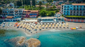 Vista aérea de un pueblo costero con una zona de playa que presenta numerosos sombrillas y tumbonas, un edificio con el nombre 'LACARAVELLA', y un fondo de edificios residenciales y vegetación.