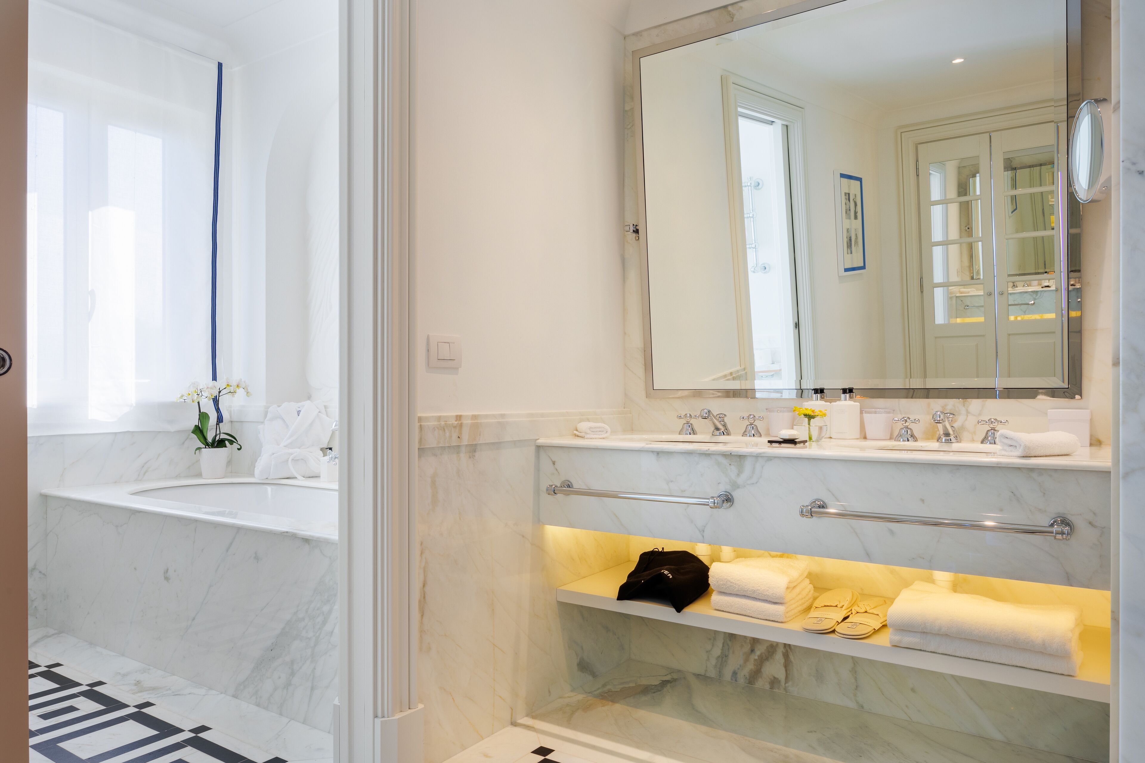 A modern bathroom with a large mirror, marble countertops, and a built-in shelf with towels and toiletries.