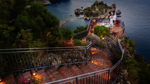 Una terraza costera con una escalera de piedra, plantas en macetas y vistas al mar y afloramientos rocosos.