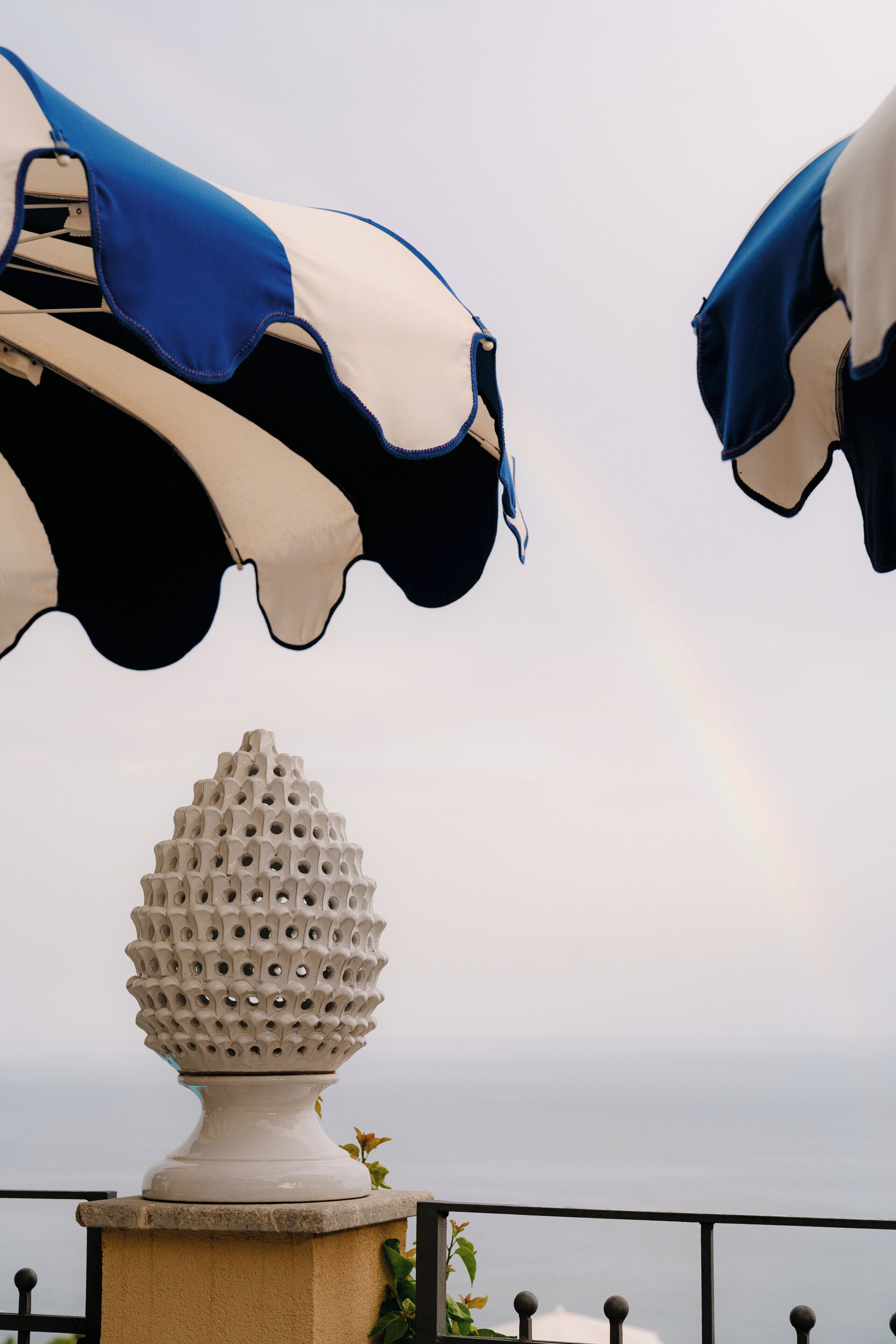 Une urne décorative blanche posée sur un piédestal, un parasol à rayures noires et blanches et une clôture en métal.