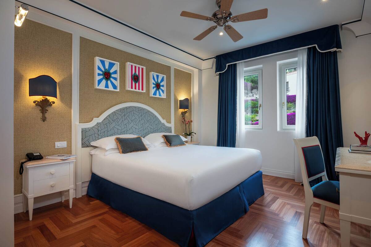 Habitació d'hotel amb un llit gran, una taula de treball amb una cadira, un ventilador de sostre i obres d'art a les parets.