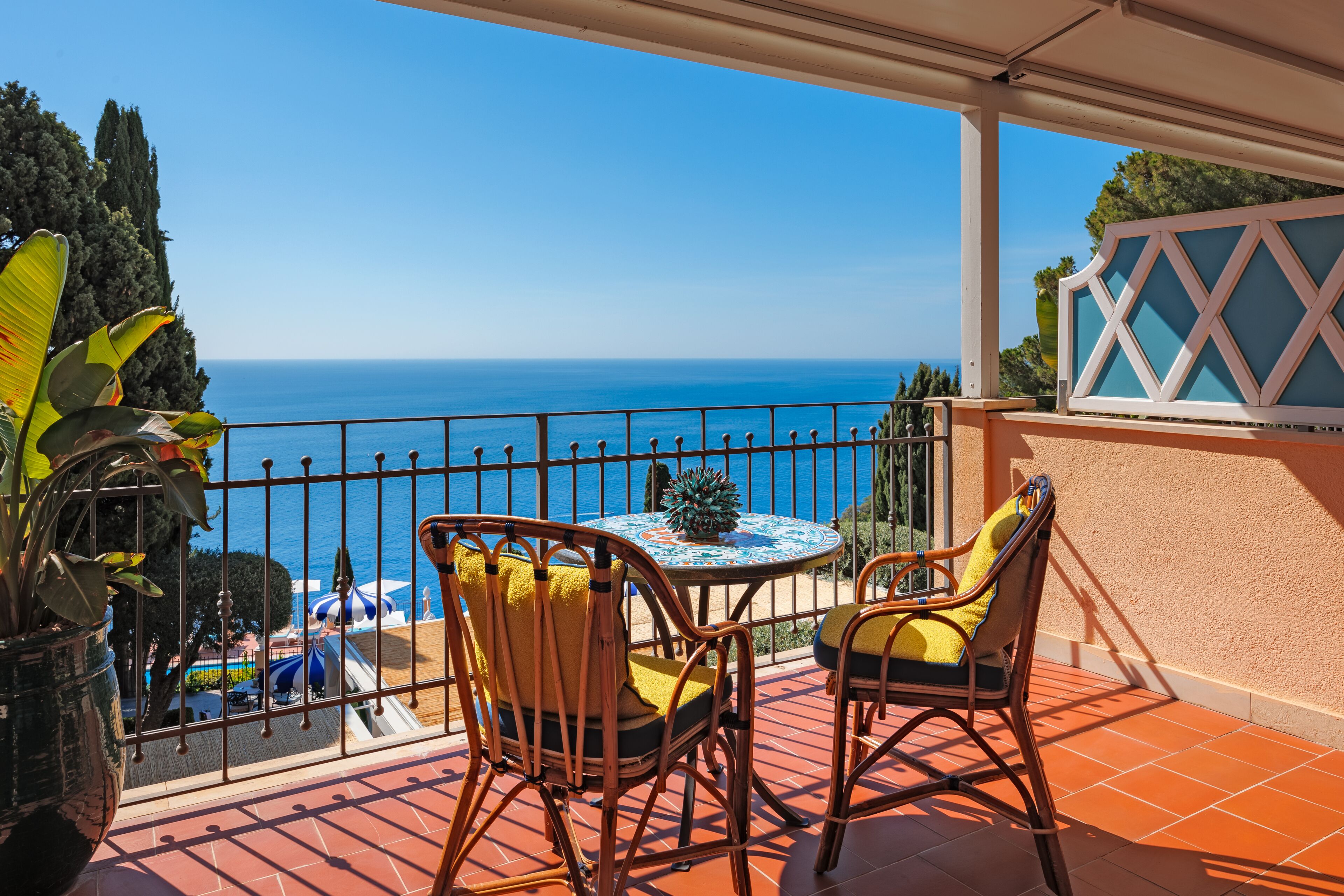 A balcony with wicker chairs and a table, overlooking a clear blue sea and a pool area.