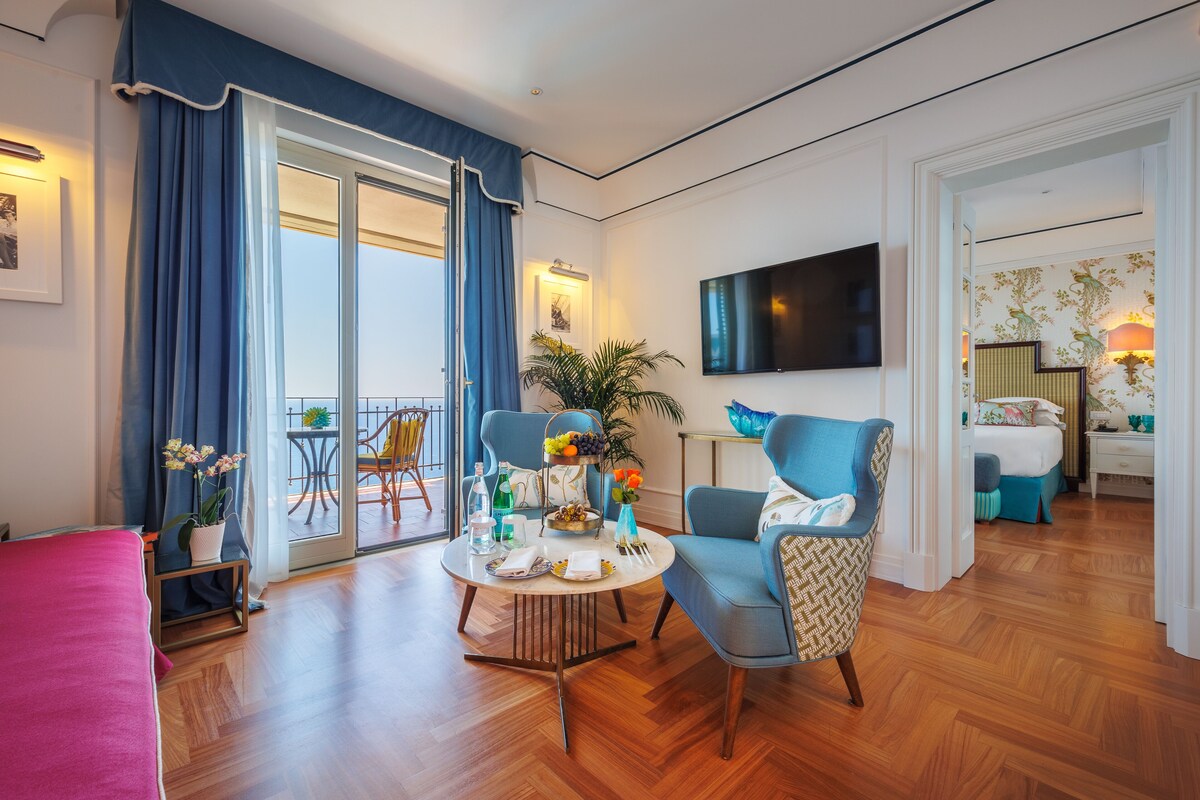Una sala de estar moderna con una mesa redonda, sillones azules, un televisor de pantalla plana y un balcón con cortinas azules.