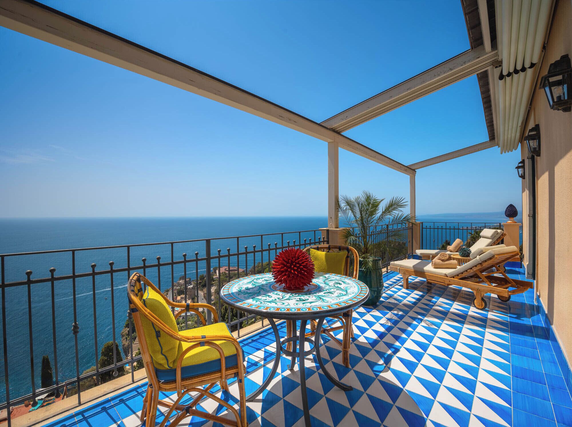 A balcony with a blue and white checkered floor, a round table with a red centerpiece, yellow chairs, and a view of the sea.