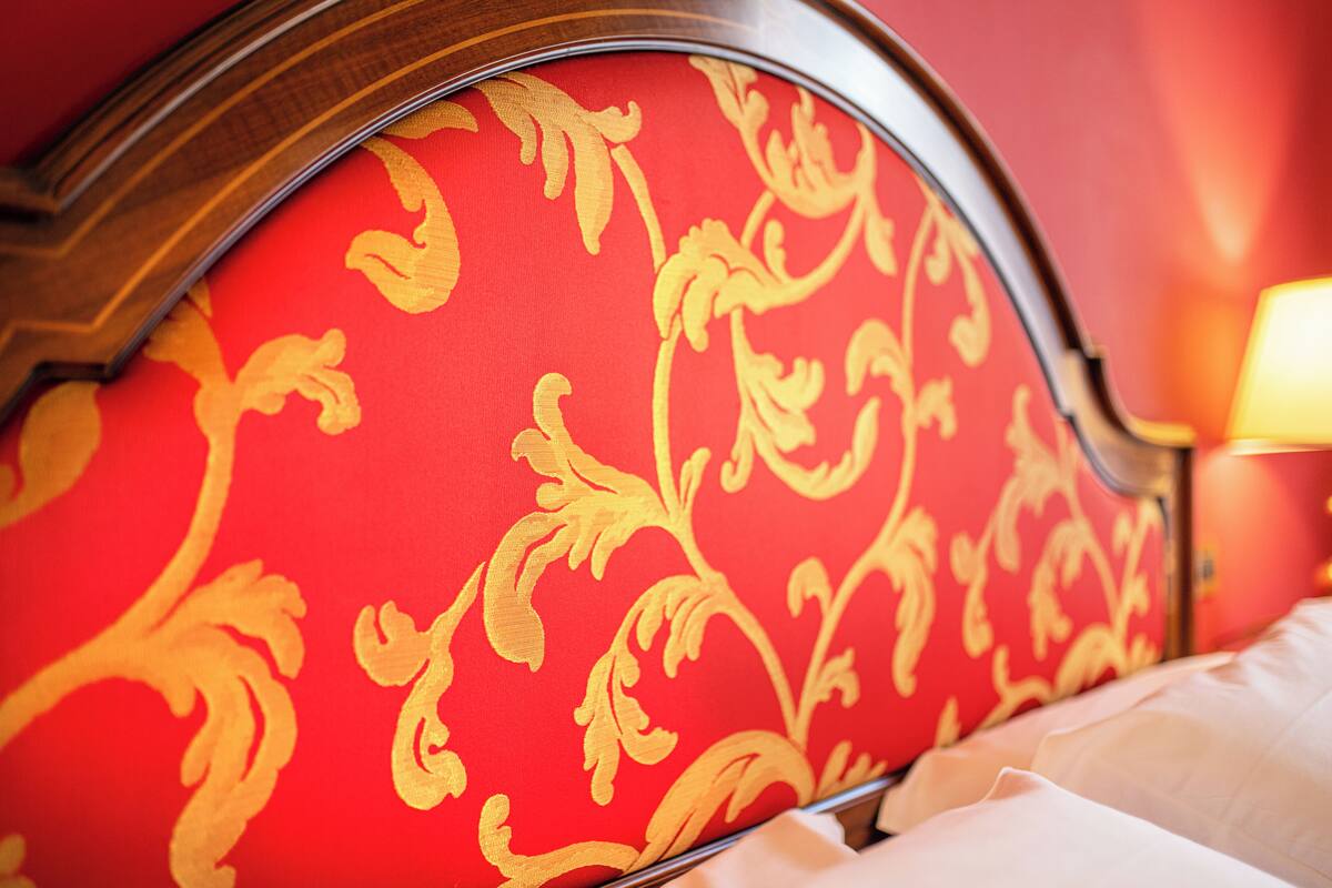 A close-up of a red upholstered headboard with a gold pattern, a bed with white linens, and a lamp on the side.
