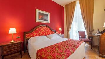 Ein Hotelzimmer mit roter Wand, einem Bett mit roter und goldener Bettdecke, einem Nachttisch aus Holz mit Lampe, einem Schreibtisch mit Stuhl und einem Fenster mit Vorhängen.