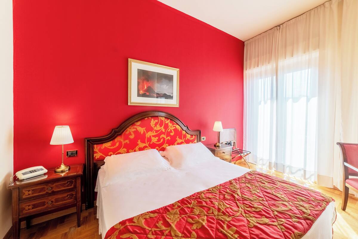 A hotel room with a red wall, a bed with a red and gold patterned bedspread, two bedside tables with lamps, a framed picture on the wall, and a window with curtains.