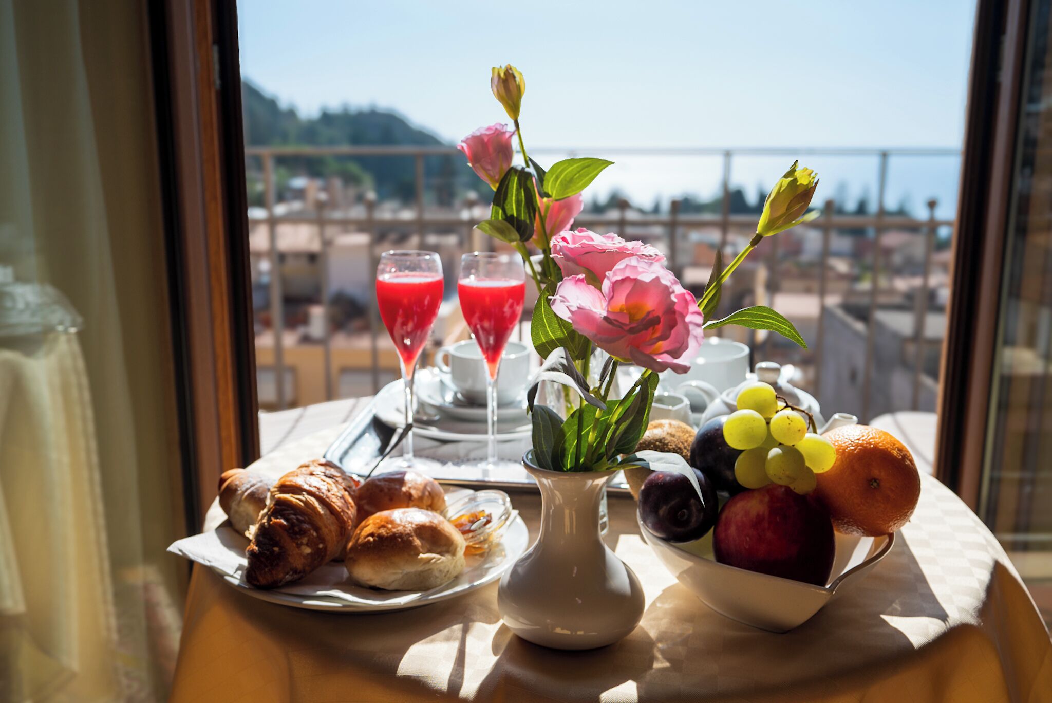 A table with croissants, a fruit bowl, a vase with flowers, and two glasses of red juice.
