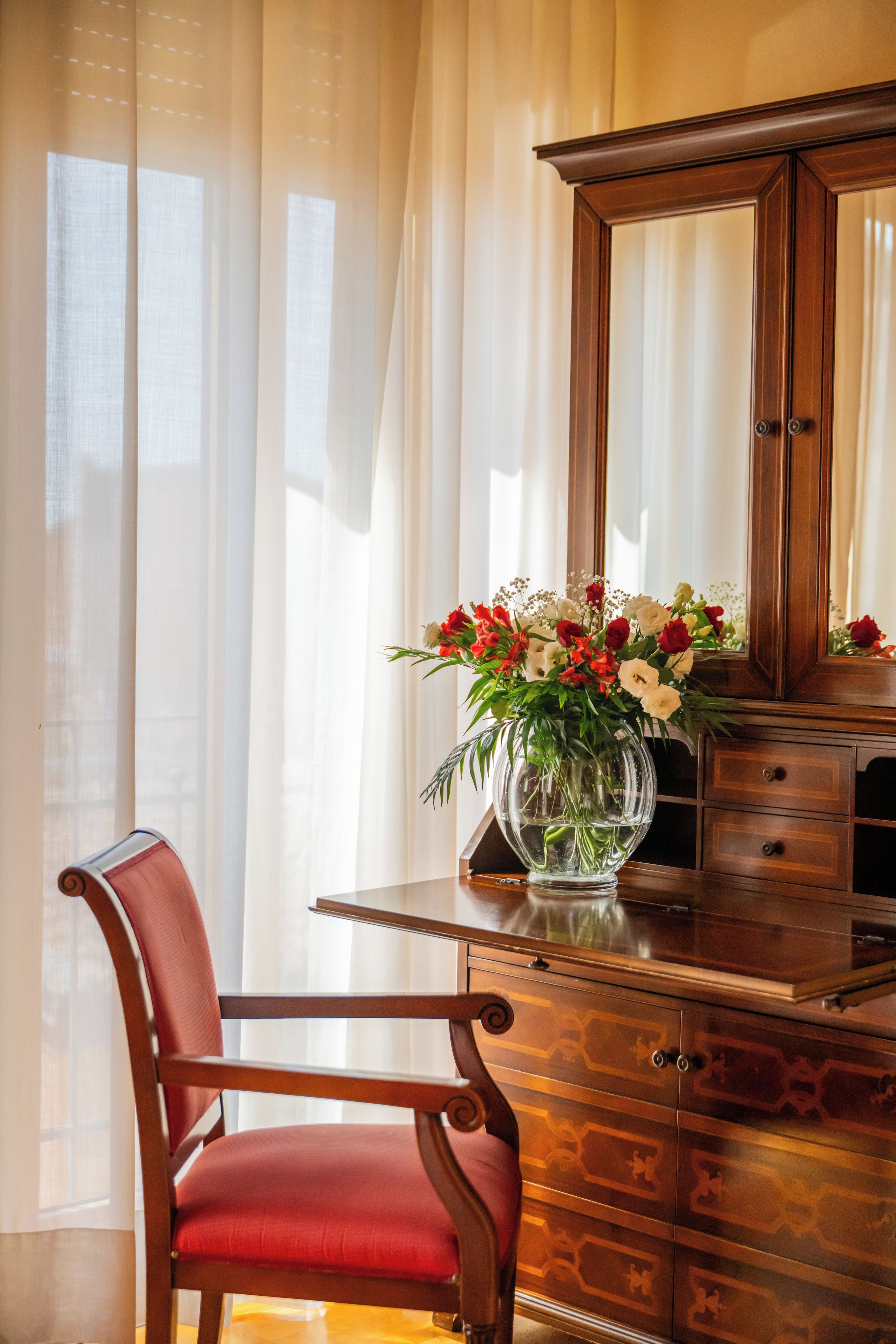 A wooden desk with a vase of flowers, a chair, and a mirror.