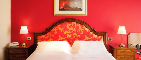 A hotel room with a red wall, a framed picture of a volcano, a wooden headboard, two bedside tables with lamps, and a bed with a red and gold patterned bedspread.
